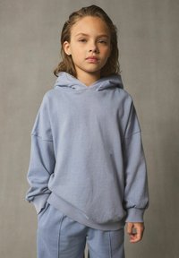 Hoodie azzurro chiaro realizzato in tessuto morbido, con una vestibilità ampia, maniche larghe e una tasca frontale. Abbinato a pantaloni azzurri chiari coordinati.