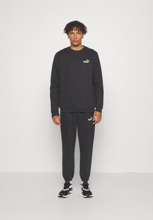 Sudadera negra y joggers a juego hechos de una tela suave. Presenta un pequeño logo blanco en el pecho y el muslo. Las zapatillas son negras con acentos rojos.