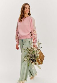 Růžová mikina s květinovými rukávy, spárovaná se světle zelenými širokými kalhotami. Modelka drží tkaný koš s květinami.