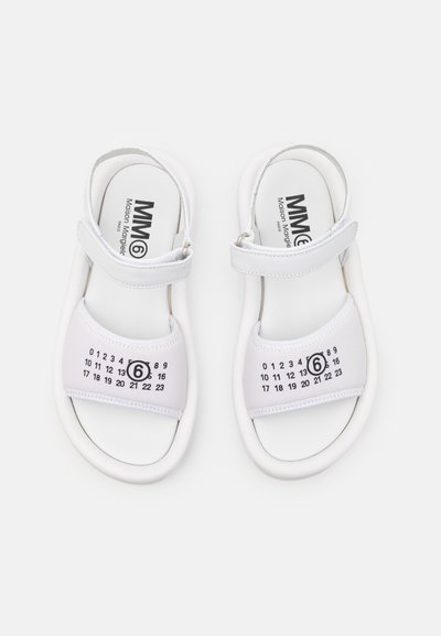 Hvite sandaler med en blank finish, med en stropp over foten og et kalendertrykk på oversiden. Myk, glatt tekstur, minimalistisk design.