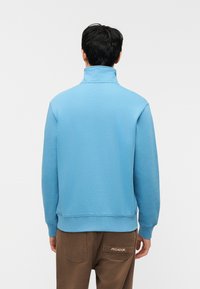 Άνδρας που κοιτάζει προς τα πίσω φοράει γαλάζιο φούτερ με ψηλό γιακά και καφέ παντελόνι με πίσω τσέπη που φέρει την ετικέτα «PEGADOR» σε λευκό φόντο.