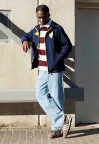 Mörkblå vindjacka med gula detaljer, randig bordeaux och vit skjorta, ljusblå jeans och bruna sneakers med vita ränder. Stående pose.