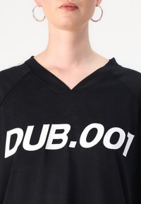 Melnā V veida krekls ar izteiksmīgām baltām burtiem "DUB.001." Izgatavots no mīksta auduma ar brīvu piegriezumu, papildināts ar sportisku dizainu.