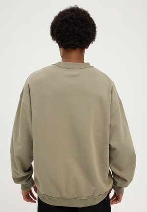 Mand med kort krøllet hår iført oversized olivengrøn sweatshirt set bagfra mod en ensfarvet baggrund.