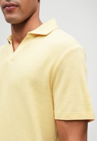 Πόλο μπλούζα σε ανοιχτό κίτρινο, υφασμάτινη πλεκτή ύφανση. Διαθέτει γιακά V και κοντά μανίκια με ίσιες άκρες. Απλός, casual σχεδιασμός.
