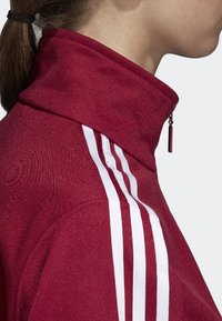 Roter Sweatshirt mit hohem Kragen und Reißverschluss, der drei senkrechte weiße Streifen auf der linken Schulter und einen strukturierten Stoff aufweist.