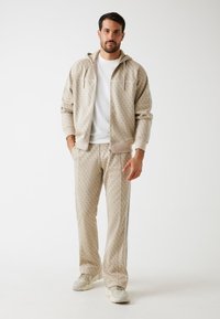 Sudadera beige con estampado y pantalones a juego con acentos blancos. Textura suave, frente con cremallera y rayas laterales en los pantalones, combinados con zapatillas gris.