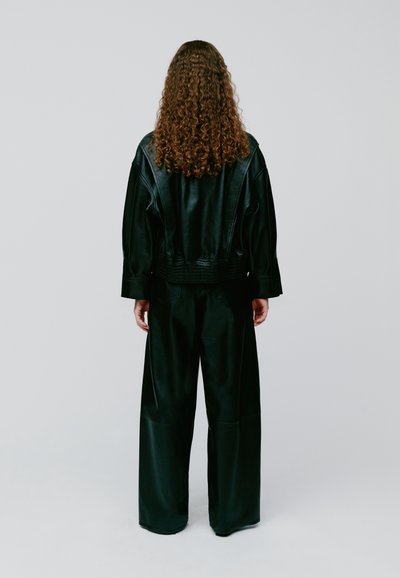 Osoba s dugom kovrčavom kosom stoji okrenuta leđima, nosi crnu kožnu jaknu i široke crne hlače na jednobojnoj pozadini.