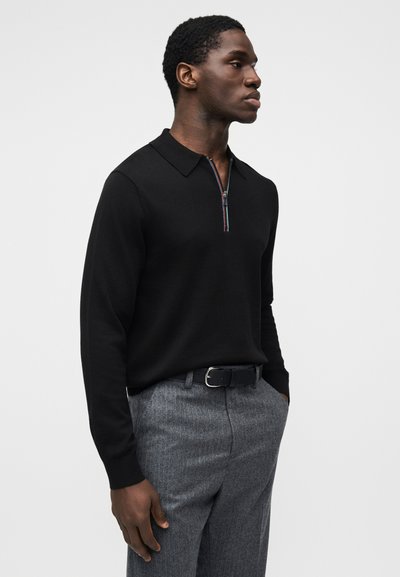 Μαύρο πόλο με μακριά μανίκια και κλείσιμο με φερμουάρ, διακοσμημένο με πολύχρωμη ρίγα σε αντίθεση. Συνδυασμένο με ανοιχτό γκρι υφασμάτινο παντελόνι και μαύρη ζώνη.