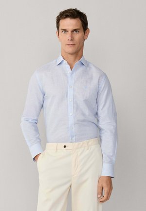 Hombre vestido con camisa de rayas azul celeste abotonada y pantalones color crema, de pie con una mano en el bolsillo contra un fondo liso.
