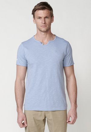 Camiseta de manga corta azul claro en una suave tela texturizada. Cuenta con un escote en V con un detalle de botón y un pequeño logo en la manga.