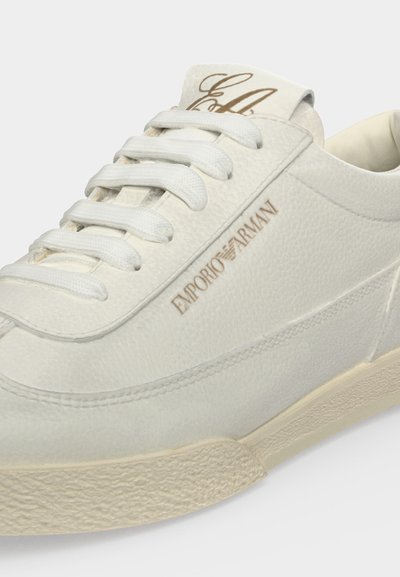 Λευκό διακοσμημένο δερμάτινο sneakers με λευκά κορδόνια, μπεζ σόλα και λογότυπο Emporio Armani σε ανοιχτό καφέ στην πλευρά.