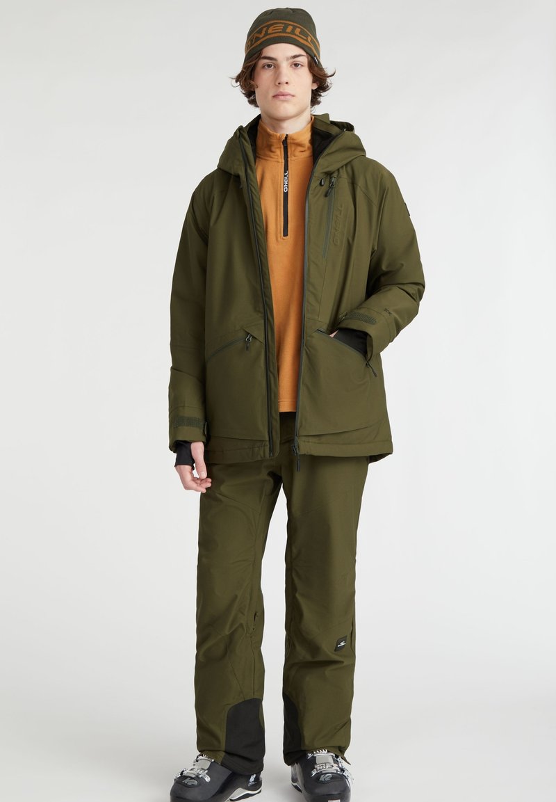 Olivgrüne Skijacke mit Kapuze, Reißverschlusstaschen und Logodetails, kombiniert mit passenden Hosen und schwarzen Akzenten. Strukturierter Stoff.