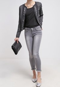 Szara dżinsowa kurtka z dekoracyjnymi guzikami i teksturowanymi akcentami, zestawiona z dopasowanymi szarymi dżinsami i srebrnymi płaskimi butami, trzymająca czarną kopertówkę.