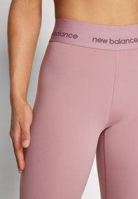 Pogled od blizu na osobu koja nosi mauve leggingse New Balance s elastičnom pojasom, ruka odmara pored kukova.