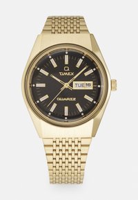 Montre à quartz dorée avec un boîtier octogonal, un cadran noir, des index lumineux en métal, un affichage de la date et un bracelet en maille texturée dorée.
