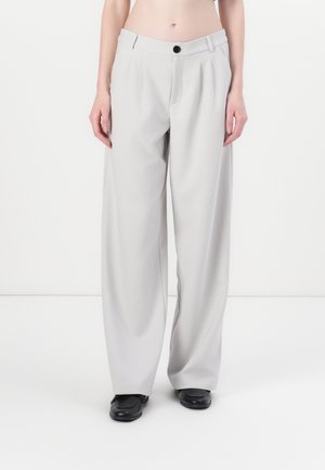 Osoba ubrana w jasnoszare spodnie o wysokim stanie i szerokich nogawkach oraz czarne buty z kwadratowym noskiem na białym tle.