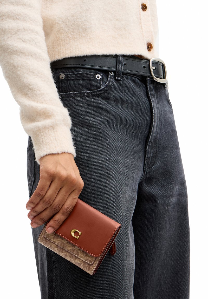Main tenant un petit portefeuille en cuir marron, portant un pull crème et un jean noir avec une ceinture noire.
