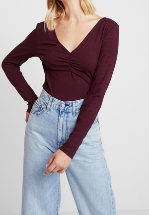 Vrouw met een geribd, lang mouws, diep V-hals bordeauxrood topje en hoge taille lichtblauwe spijkerbroek, met één hand op haar heup.