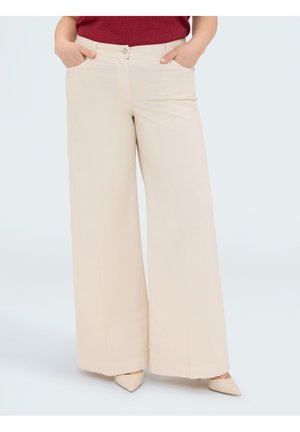 Femme portant un pantalon beige à jambes larges avec poches avant, associé à des chaussures beiges pointues et un haut rouge, debout les mains dans les poches.