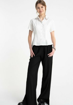 Młoda kobieta stoi z rękami w kieszeniach czarnych spodni o szerokich nogawkach, ubrana w dopasowaną białą, krótkorękawową koszulę na guziki i czarne buty.