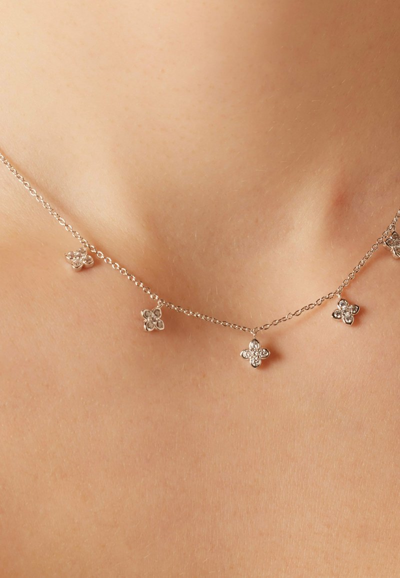 Zilveren halsketting met kleine bloemenvormige hangers, sommige versierd met stenen, aan een delicate ketting. Zachte textuur met een glanzende afwerking.