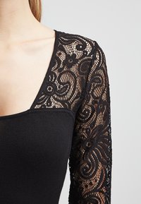 Černý top s přiléhavým tělem ze látky a dlouhými průhlednými krajkovými rukávy s propracovanými květinovými vzory. V-řez a hladká textura.
