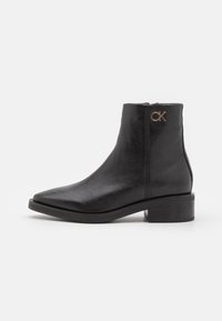 Bottine en cuir noir avec bout pointu, talon carré, fermeture éclair latérale et accent logo doré sur le côté extérieur. La texture semble lisse.