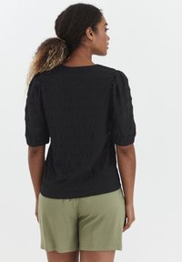 Blusa nera con texture e maniche corte a sbuffo, abbinata a pantaloni corti verde oliva. Il tessuto presenta un motivo ondulato e una vestibilità comoda.