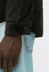 Sötét kordbársony zakómandzsetta texturált, függőleges bordákkal és fekete gomb részlettel, világoskék farmer anyaggal párosítva.