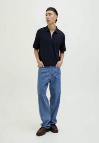 Camisa polo azul-marinho com fecho e relaxada, jeans claros de corte solto e sapatos castanhos. Design minimalista com um look casual, de pé contra um fundo neutro.