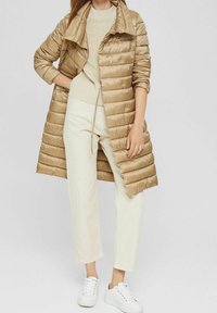 Beige Daunenjacke mit hochgeschlossenem Kragen und horizontalem Steppmuster, getragen über einem hellen Pullover und cremefarbenen Hosen, kombiniert mit weißen Sneakers.