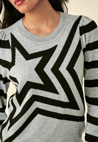 Pull grijs avec un motif d'étoiles en zigzag noir et des rayures. Texture tricotée avec un col rond et des détails froncés sur les épaules.