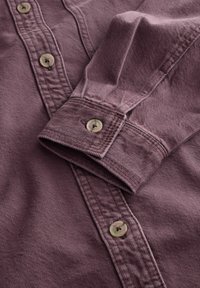 Dugave košulja dugih rukava od mauve pamučnog materijala s teksturiranim završetkom. Ima smeđe gumbe i detalj manžetni na gumb.