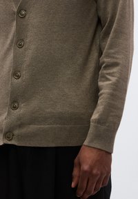 Πράσινο-καφέ ζακέτα με V-λαιμόκοψη, ριμπ μανσέτες και έξι στρογγυλά κουμπιά κατά μήκος του μπροστινού μέρους. Μαλακή πλεκτή υφή, φορεμένη πάνω από μαύρο ρούχο.