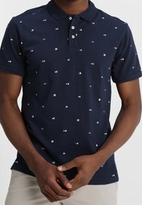 Polo bleu marine en coton, doté d'un col à boutons et de motifs géométriques blancs éparpillés pour le détail. Manches courtes, coupe standard.