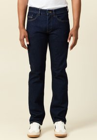 Jeans in denim blu scuro con taglio dritto, caratterizzati da un classico design a cinque tasche e cuciture a contrasto. Indossati con sneakers bianche.