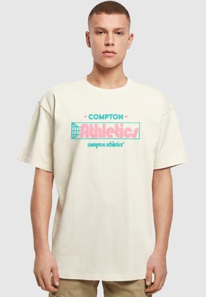 Beige oversized t-shirt lavet af bomuld med rund hals; har grafisk tekst "COMPTON Athletics" i pink og turkis.