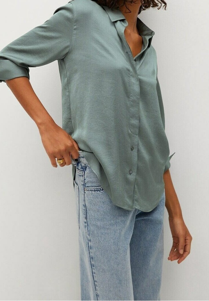 Femme portant une chemise ample vert sauge à boutons avec manches retroussées et un jean taille haute bleu clair, ajustant l'ourlet de la chemise de la main gauche.