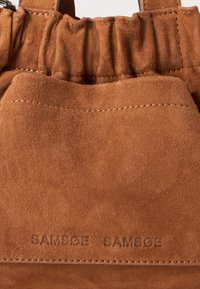 Bruine suède tas met een gathered bovenkant, geëmbosseerd "SAMSØE SAMSØE" logo en zichtbare stiksels. Zachte textuur, compacte vorm.