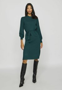 Rochie lungă, de culoare verde închis, cu mâneci lungi, evidențiată de un cordon la talie și o fanta laterală. Material tricotat cu o textură netedă, asortată cu cizme negre până la genunchi.