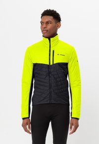 Uomo che indossa una giacca a zip gialla e nera e pantaloni neri, mentre guarda di lato su uno sfondo bianco semplice.