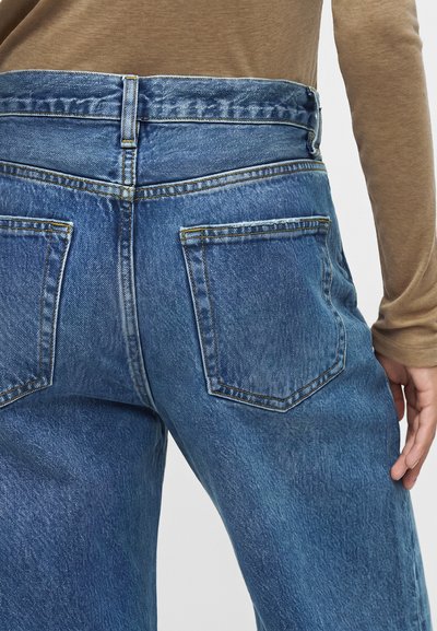 Personne portant un jean bleu en denim à coupe ample et un haut beige à manches longues, vue de dos à la taille et aux hanches.