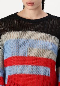 Pull en tricot avec un tissage lâche et ouvert dans des rayures noires, rouges, bleues et beiges ; col rond et design de blocs de couleurs distinct.