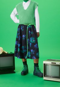 Un gilet en tricot vert sur une chemise blanche, associé à une jupe florale noire avec des accents verts et des bottes lacées vertes. Fond vert uni.