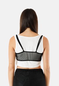 Bralette in rete nera sovrapposto a una canotta bianca. I bretelli sono regolabili e il fondo presenta un design fitted e cropped. Pantaloni neri a vita alta.