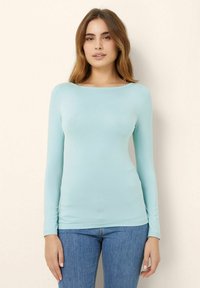 Helleblaue langärmlige Bluse aus dehnbarem Stoff mit rundem Halsausschnitt, taillierter Passform und glatter Textur, kombiniert mit Jeans.