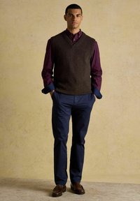 Vīrietis stāv ar rokām kabatās, ģērbies brūnā bezsoma džemperī, bordo pogājama krekla, tumši zilās biksēs un brūnās mokasīnās uz beidžas fona.