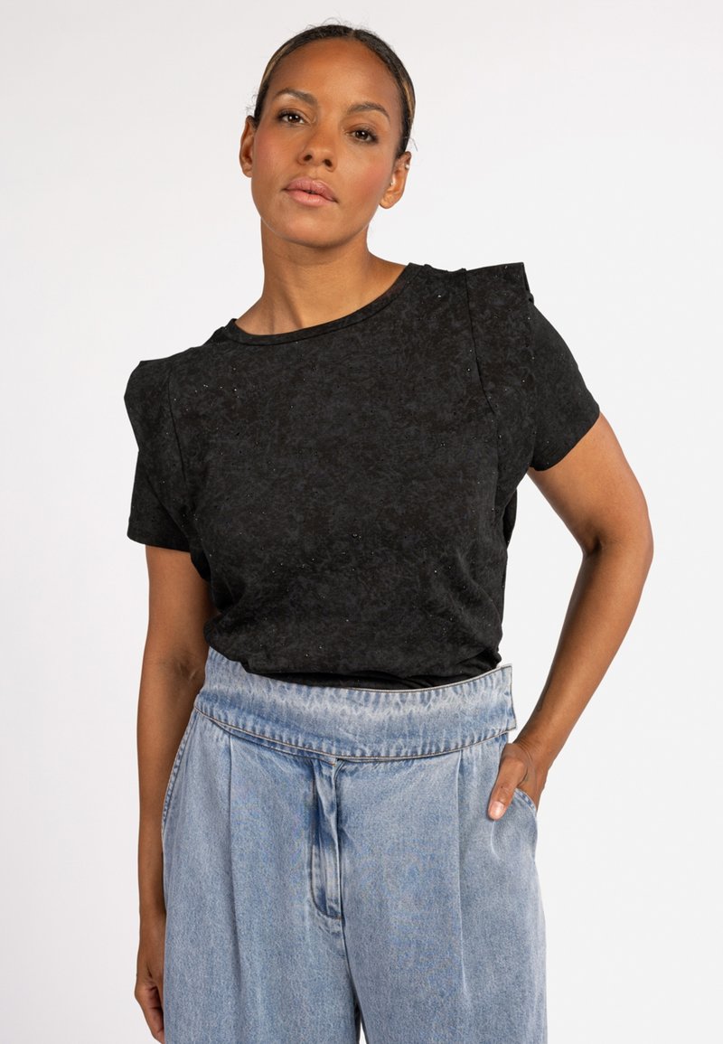 Zwarte blouse met korte mouwen, gemaakt van textiel met een subtiele glans. Heeft een ronde halslijn en schouderaccenten. Gecombineerd met lichtgekleurde spijkerbroek.