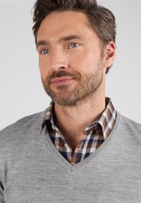 Maglione grigio a maglia con scollo a V sovrapposto a una camicia a quadri marrone e blu. I tessuti sembrano morbidi, con modelli distintivi di entrambi i capi.